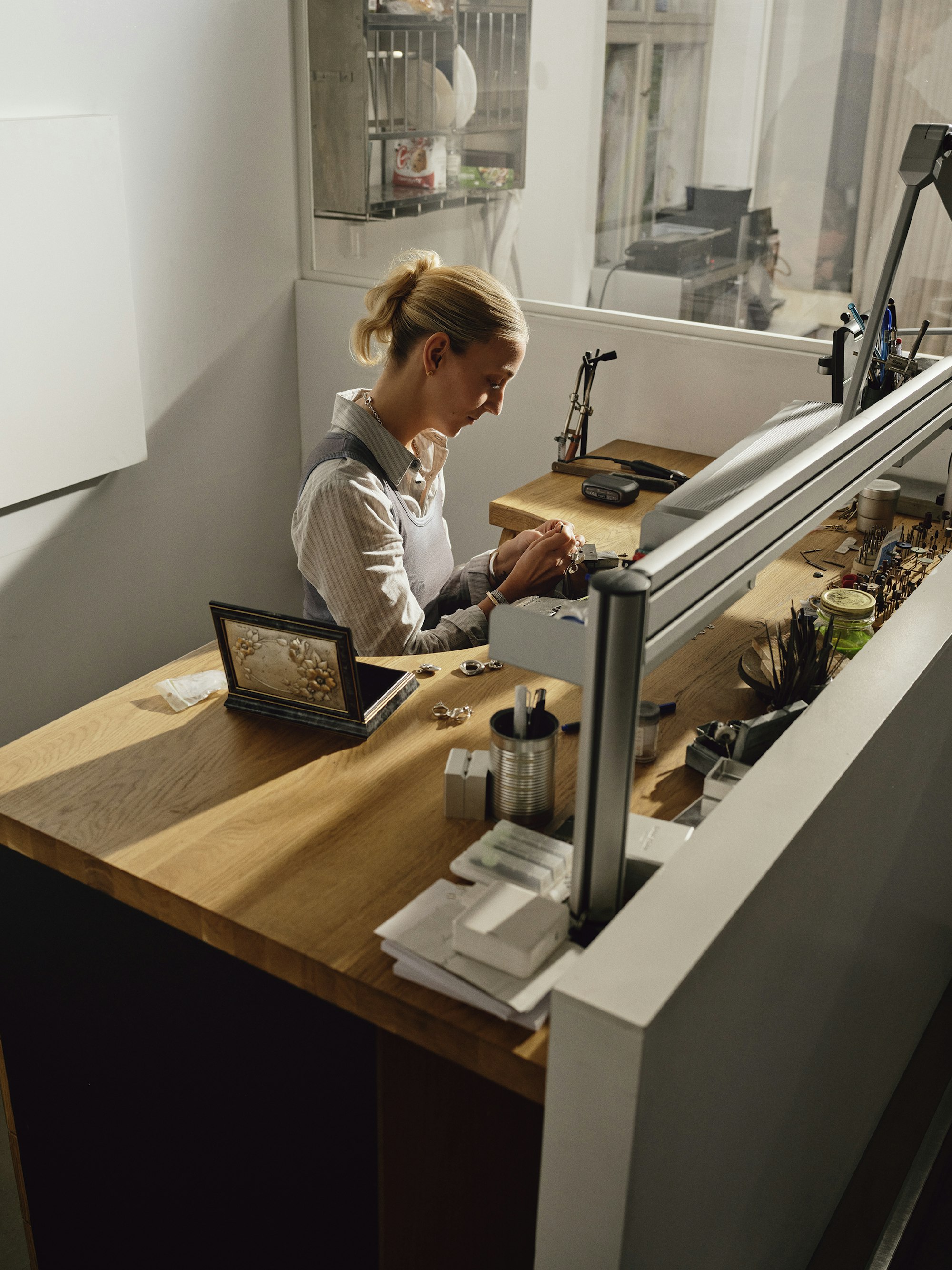 A jeweler working at her bench, focused on a piece in her hands under task lighting